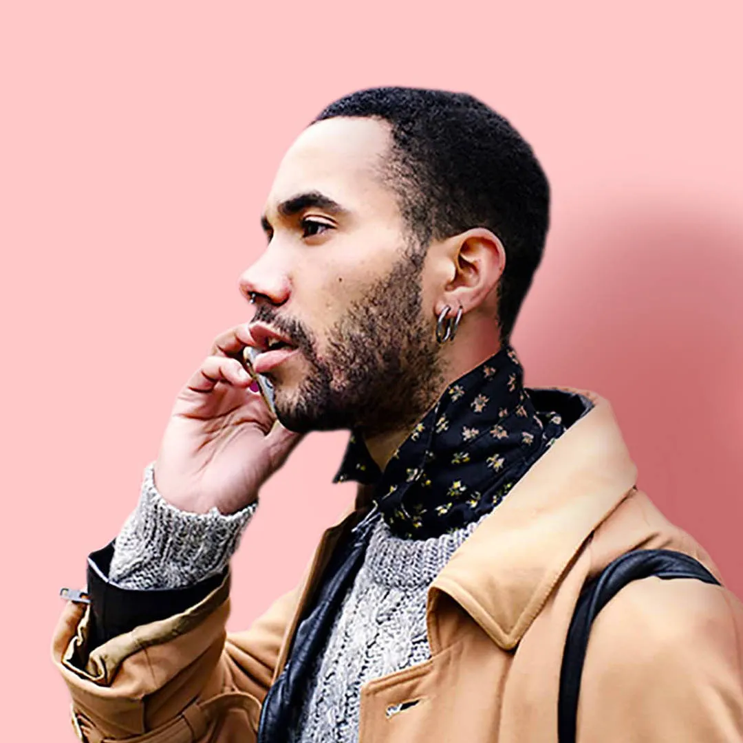 A confident and assured young man speaking on the phone. He has earrings, a patterned shirt, grey jumper and beige coat.