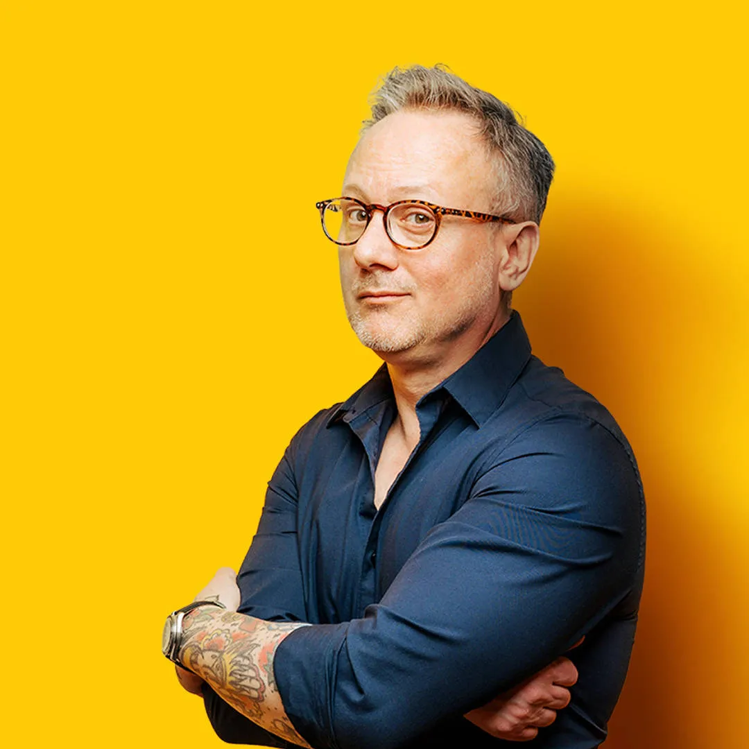 Confident man with grey hair, glasses, tattoos and a watch, wearing a navy shirt, arms crossed, looking assured and hopeful.
