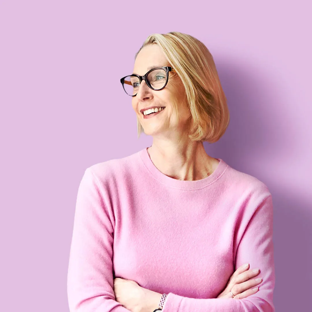 Smiling woman in her 40s with blonde bob and black glasses, wearing a pink jumper, arms crossed, looking approachable and confident.